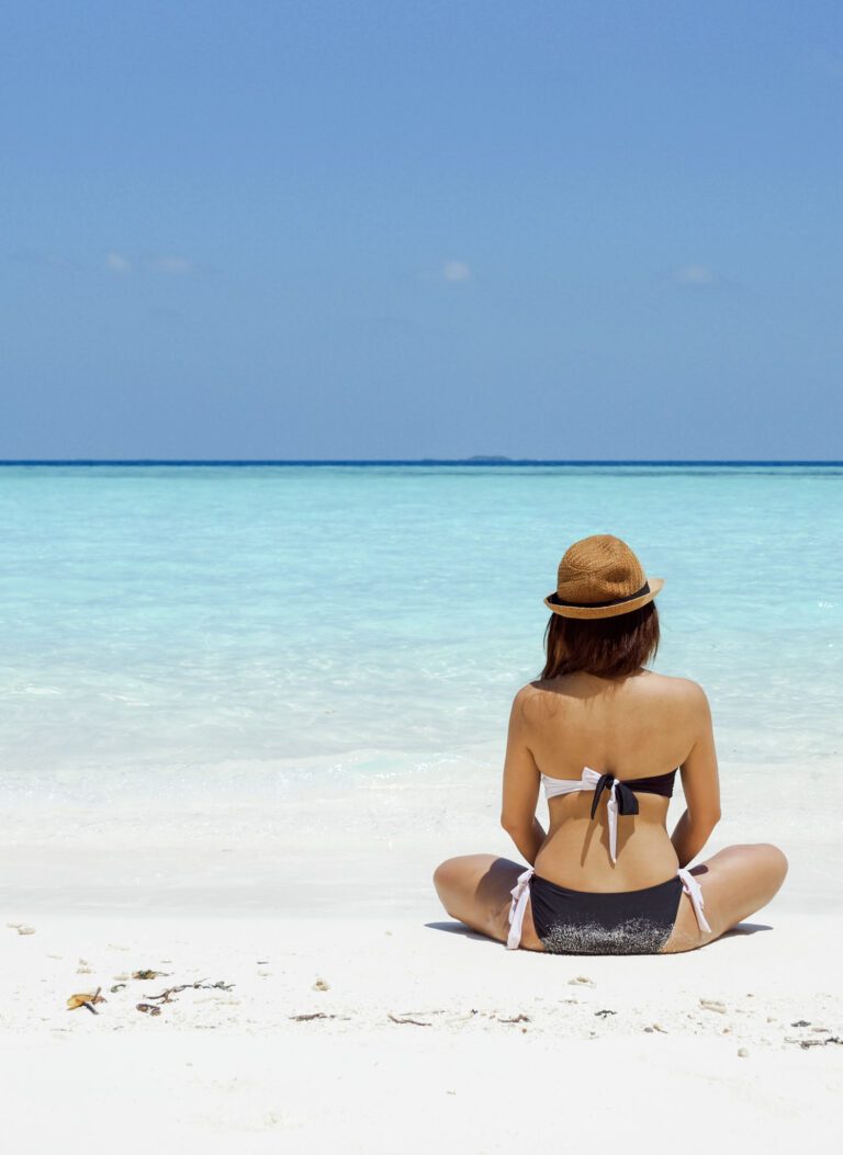 A woman sits on a white sand beach with clear blue waters. This article covers how to achieve the perfect summer glow.