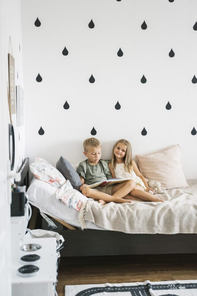 A boy and a girl read a book together on a bed. Behind them is a black and white peel and stick wallpaper. 