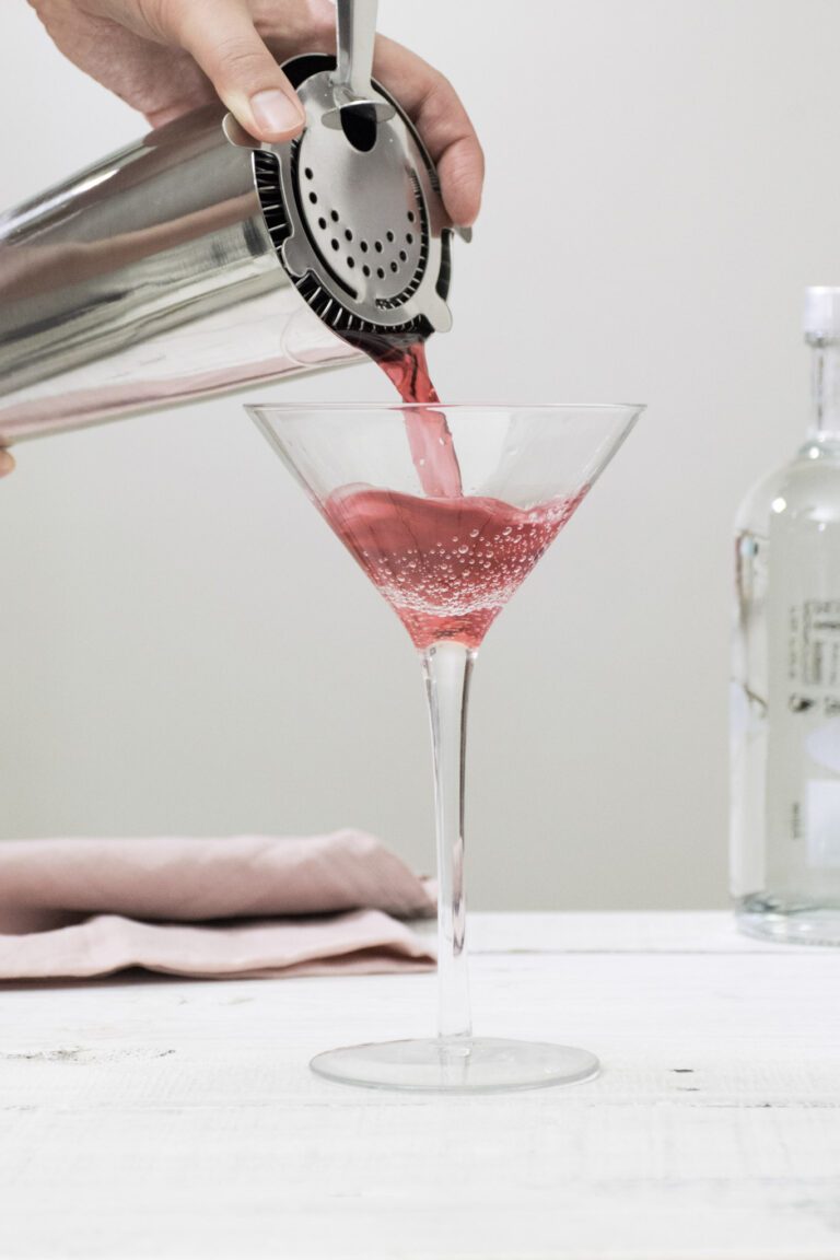 A Cosmopolitan cocktail is being mixed and poured into a glass. This article features getting to know the Cosmopolitan.