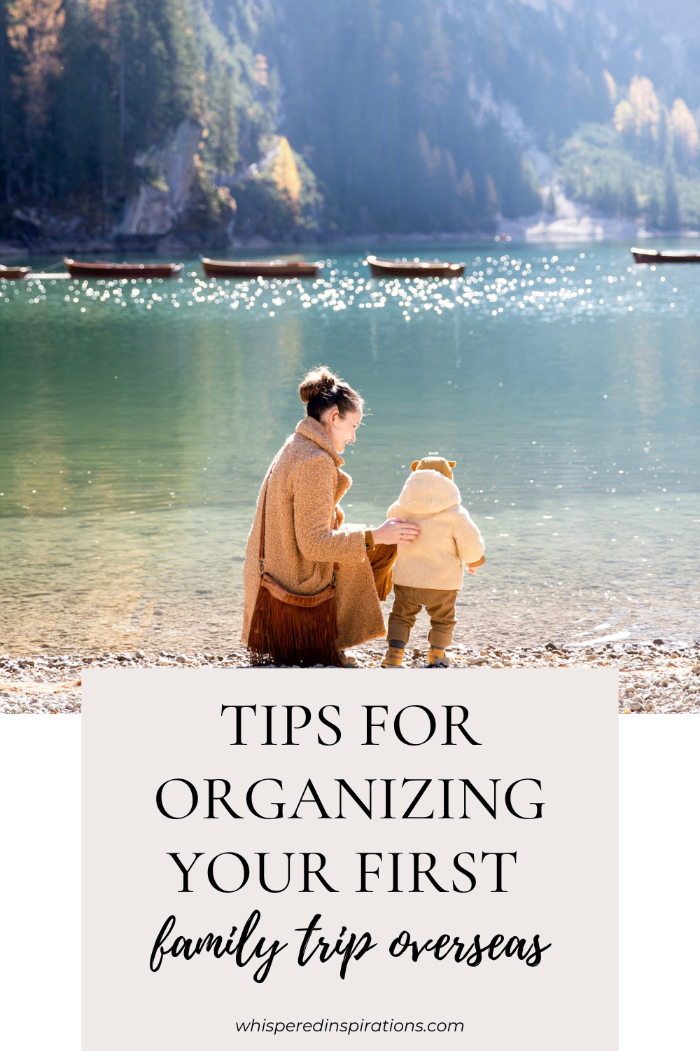 A woman kneels and holds a baby by the shore of a beautiful landscape. This article covers tips for organizing your first family trip overseas. 