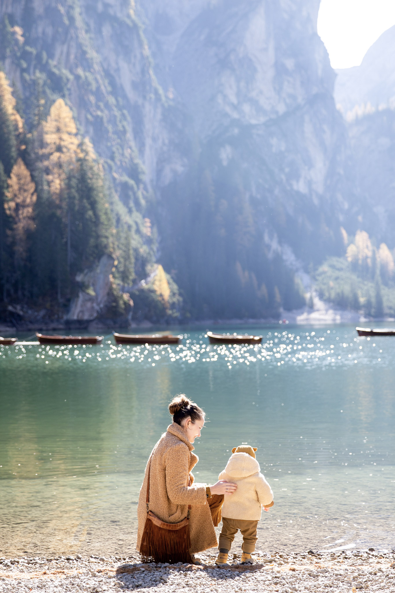 A woman kneels and holds a baby by the shore of a beautiful landscape. This article covers tips for organizing your first family trip overseas. 