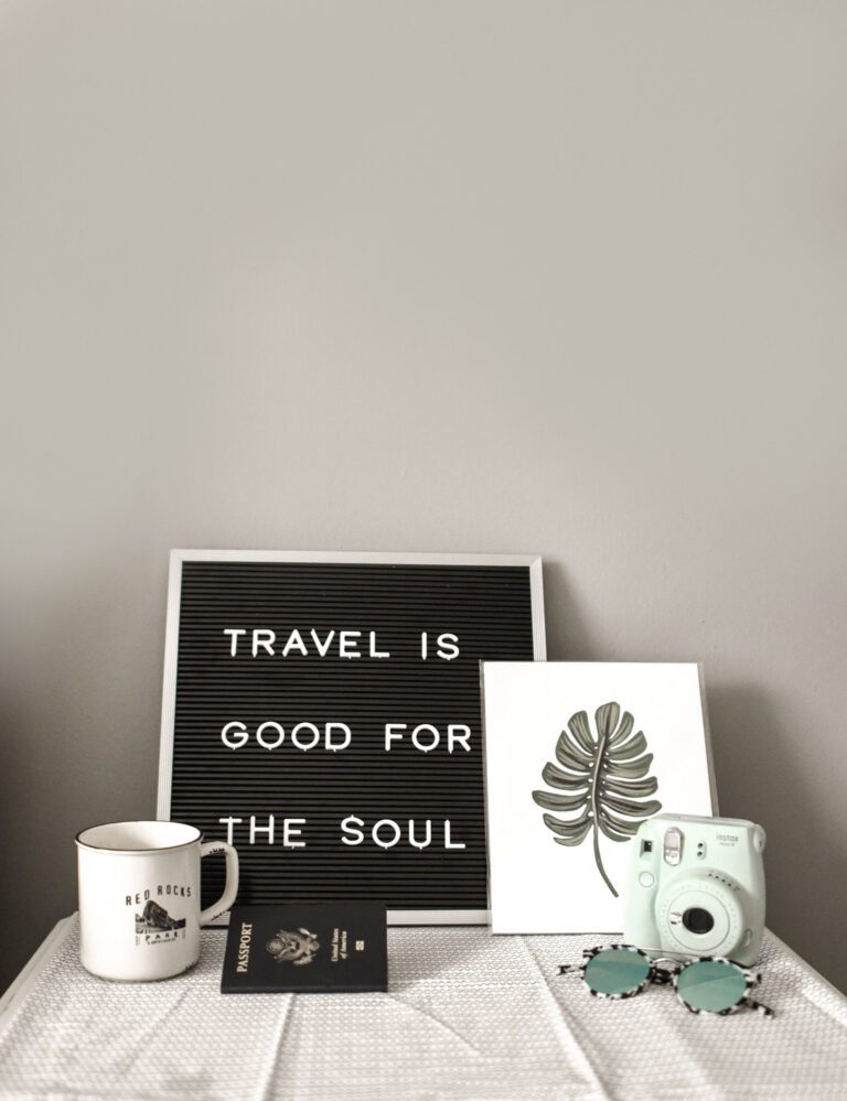A letterboard says, 'Travel is good for the soul' and is surrounded by a passport, mug, camera and sunglasses. This article covers business travel hacks that can make your life easier.