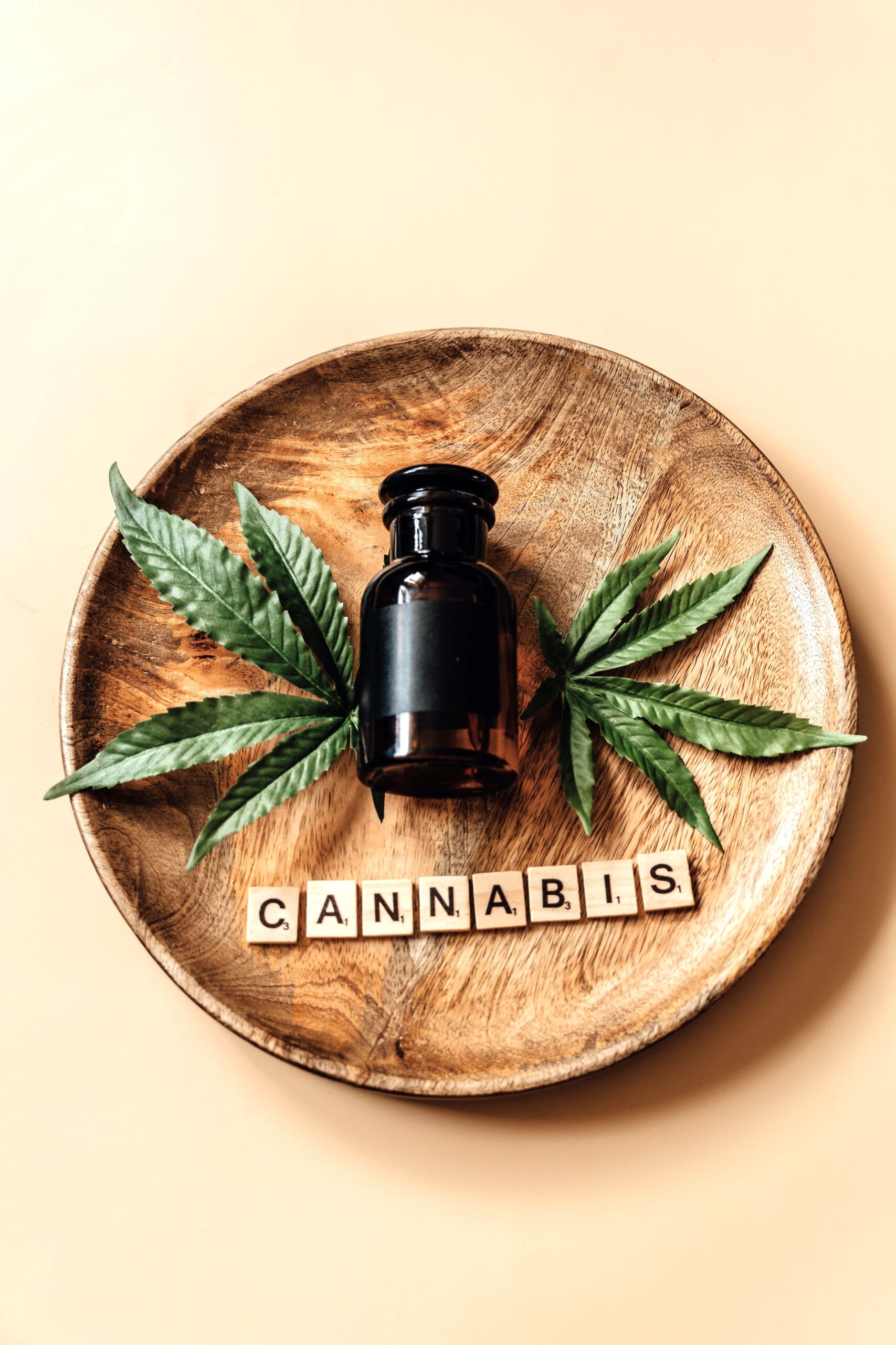 A wooden plate holds a tinted bottle on laid on top of green leaves. Scrabble letters spell the word cannabis underneath. This article covers cannabis dosing. 