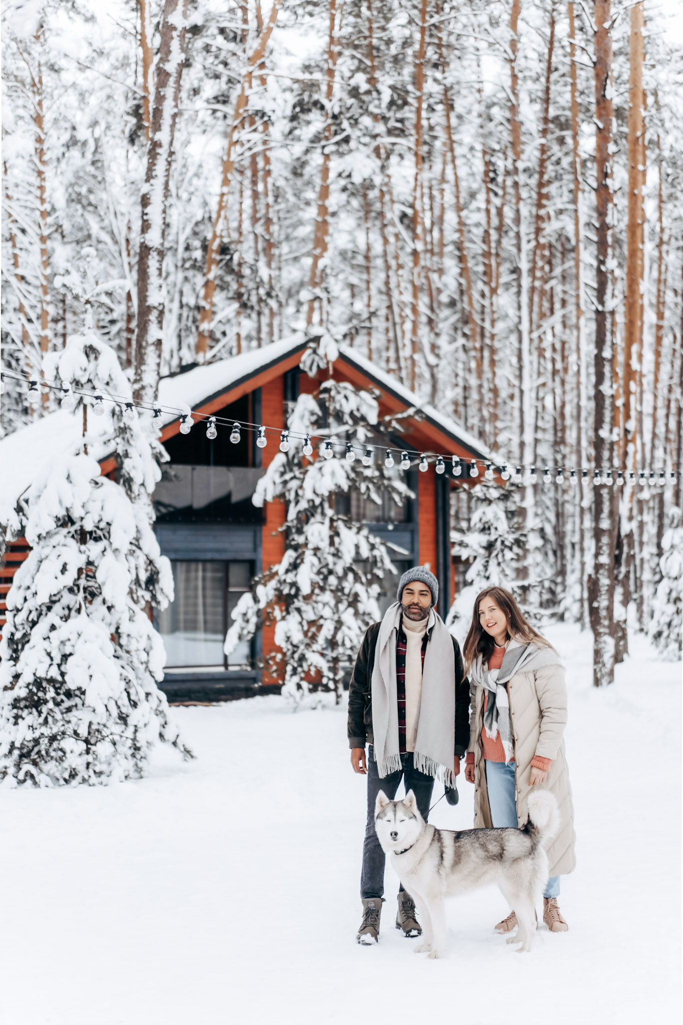 A beautiful home in a forest covered in snow. A couple with a husky are shown. This article covers creative ways to make your backyard fun and functional during the winter.