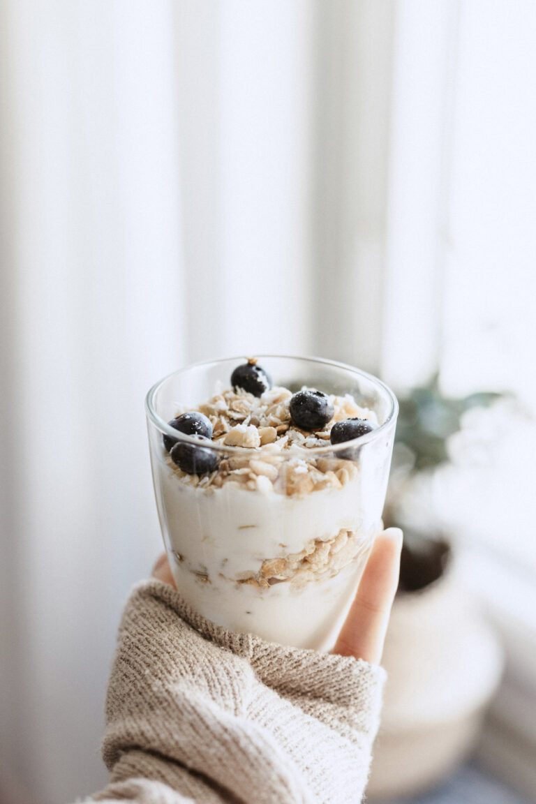 A woman wearing a brown sweater is holding up a cup of yogurt with granola and blueberries. This article covers how to make yogurt at home with a small milk pasteurizer.