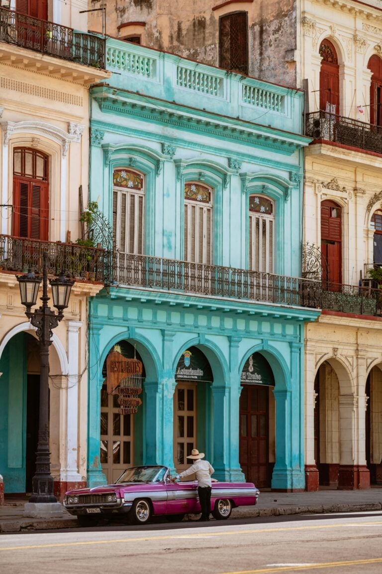 A man sits at his vintage car in Cuba, in front of the colorful buildings. This article covers facts about Cuba that make it a must-see country.