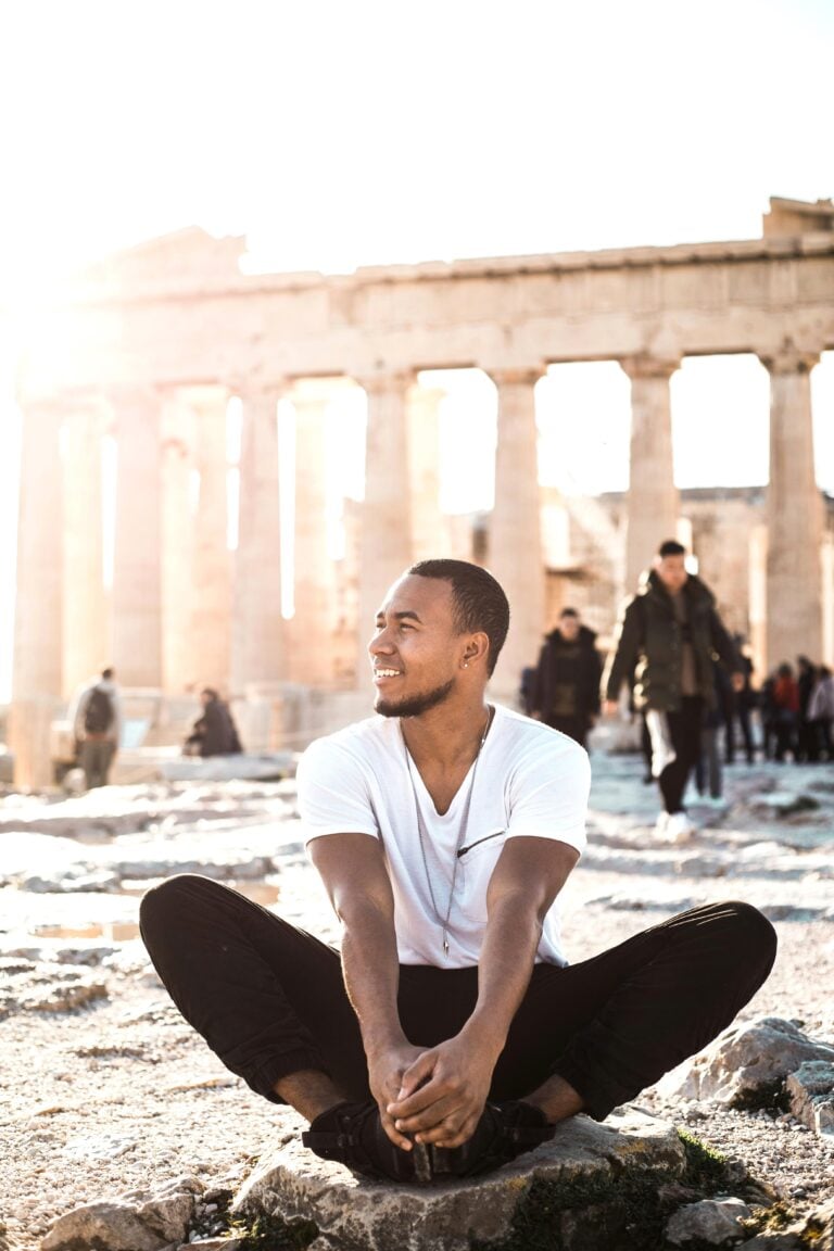 A man sits and poses in front of the Acropolis while crowds visit behind him. This article covers visiting the Acropolis in peak season and how to beat the crowds.