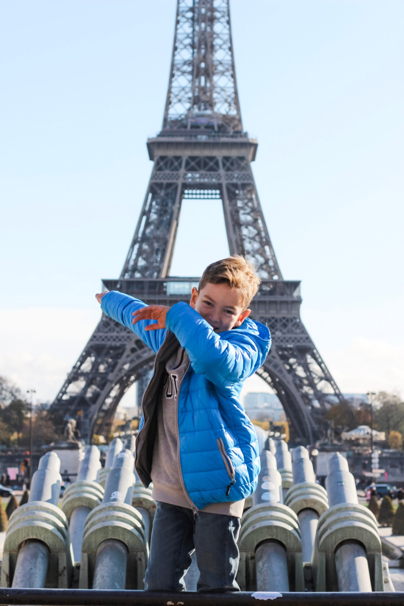 A little boy dabs in front of the Eiffel Tower. He is wearing a blue sweater. This article covers an ultimate guide to buying Eiffel Tower tickets for families with kids.
