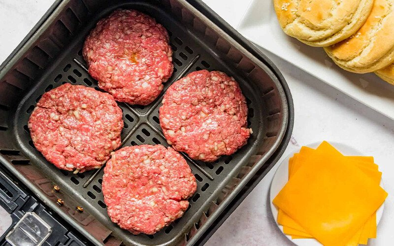 An air fryer with hamburger patties is shown. There are buns and cheese on the side. This article shares how to make the perfect air fryer hamburger.