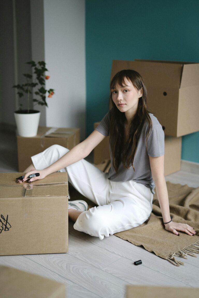 A woman sits on the ground, packing and marking boxes for moving. She stares at the camera longingly. This article covers what no one tells you about packing up and starting over.