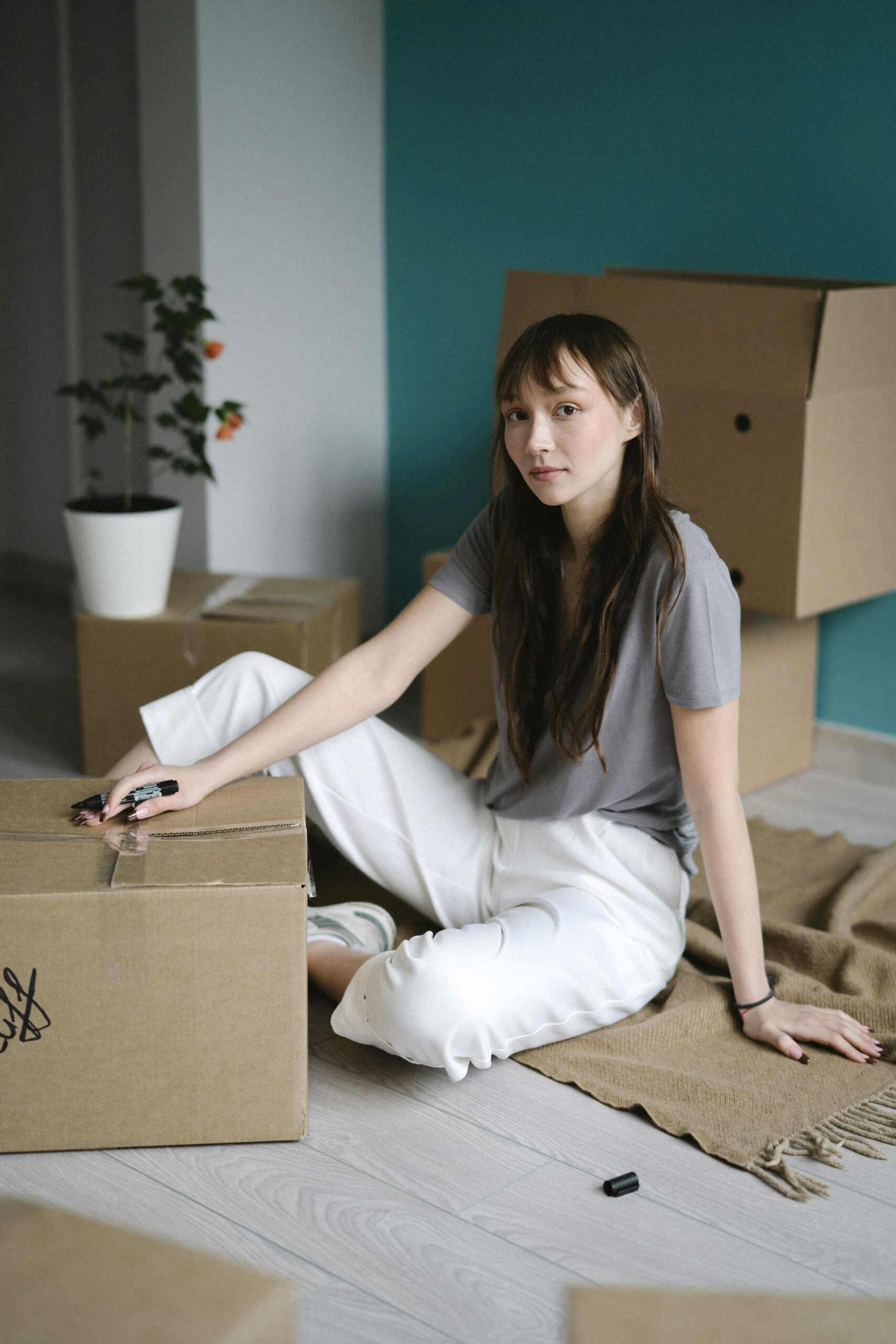 A woman sits on the ground, packing and marking boxes for moving. She stares at the camera longingly. This article covers what no one tells you about packing up and starting over.