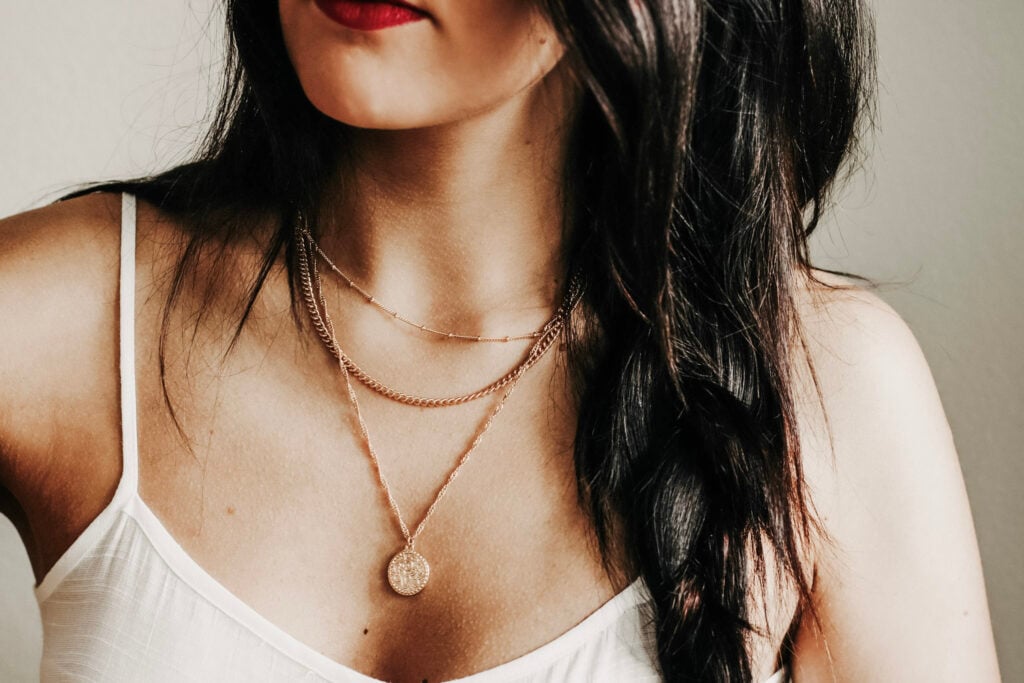 Close-up image of a young woman with dark hair wearing layered necklaces and a white top. This article covers St. Christopher Necklaces and how to choose one and style it effortlessly.