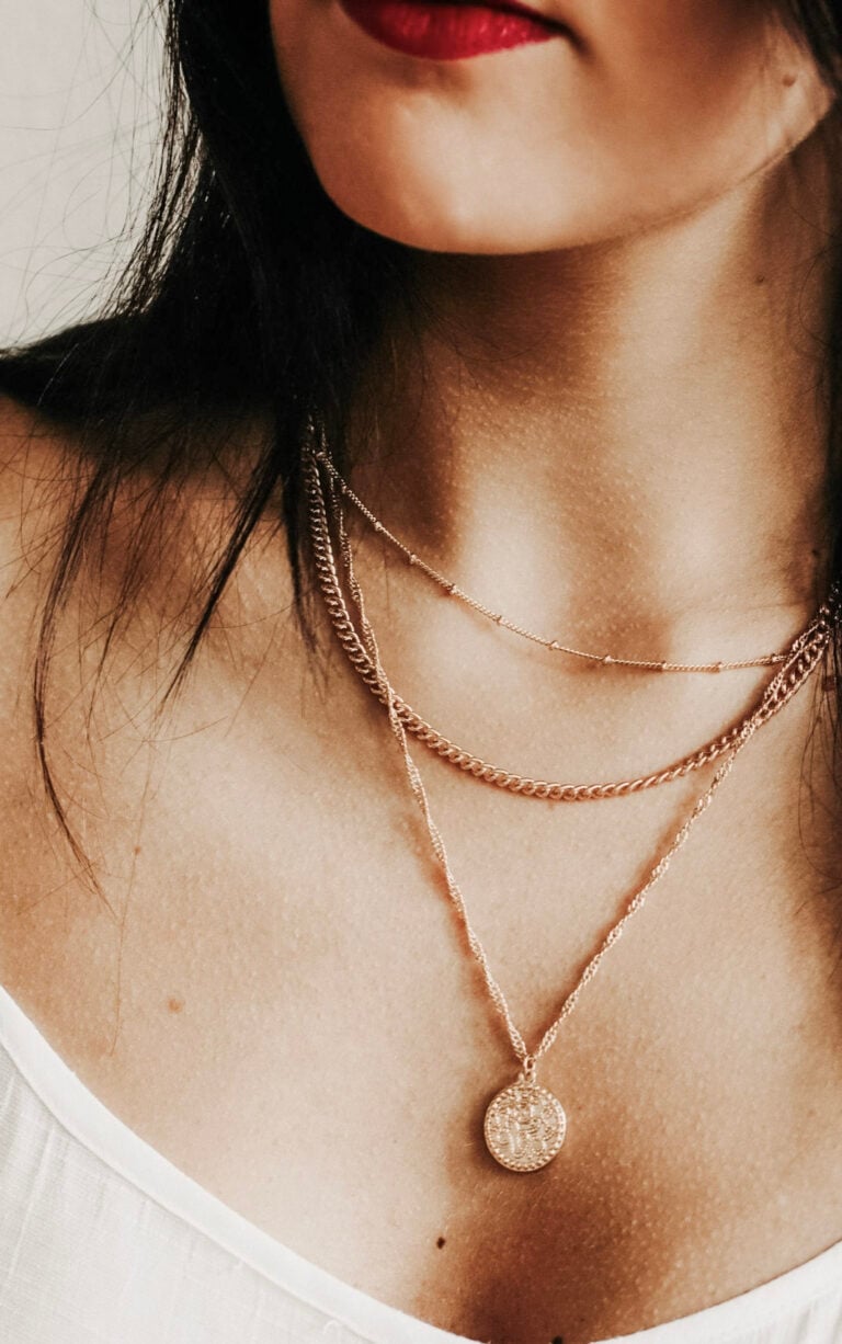 Close-up image of a young woman with dark hair wearing layered necklaces and a white top. This article covers St. Christopher Necklaces and how to choose one and style it effortlessly.