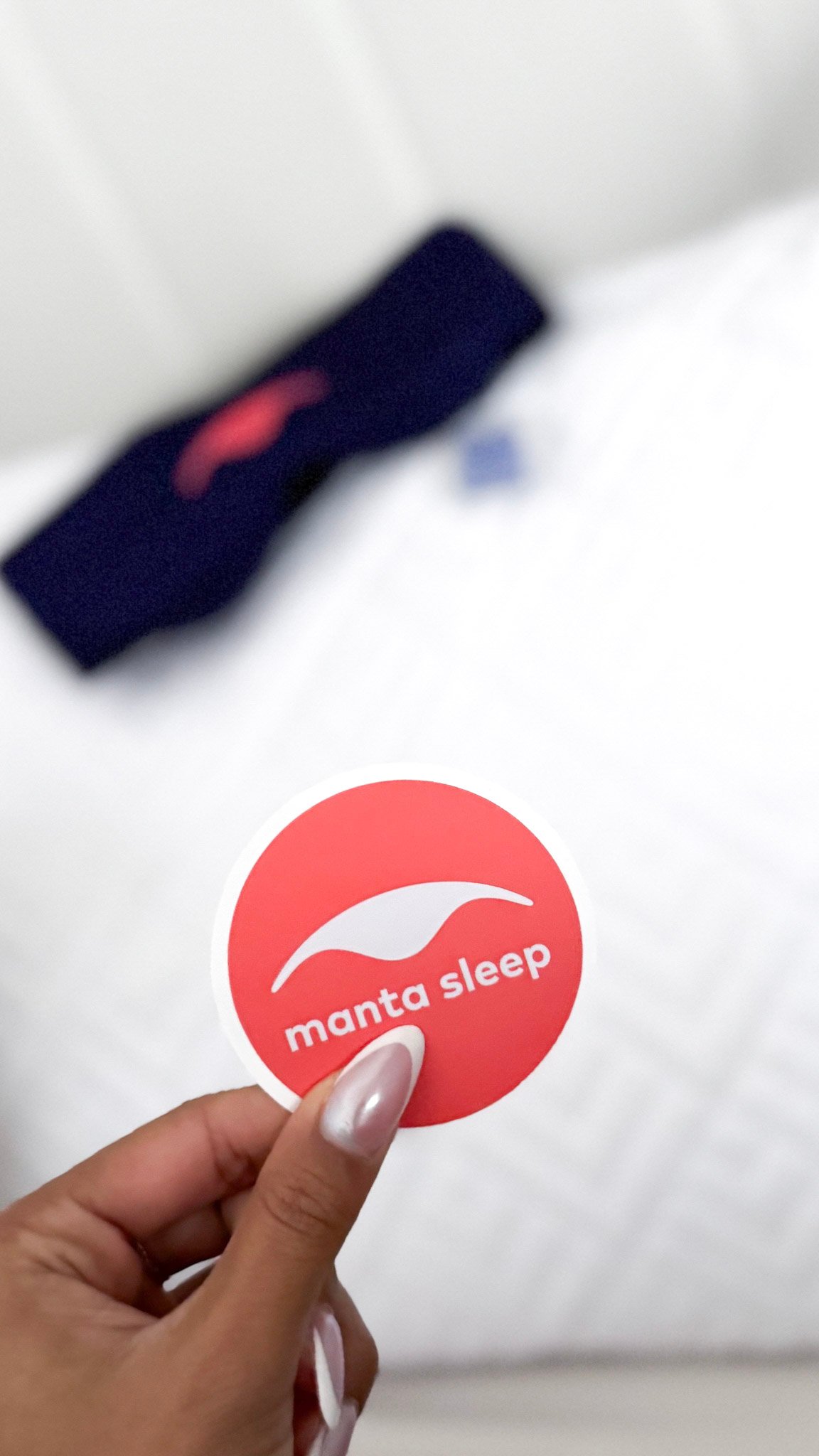 A hand holds a Manta Sleep sticker and in the background, on a white pillow, is a blurred purple Manta Silk Sleep Mask.