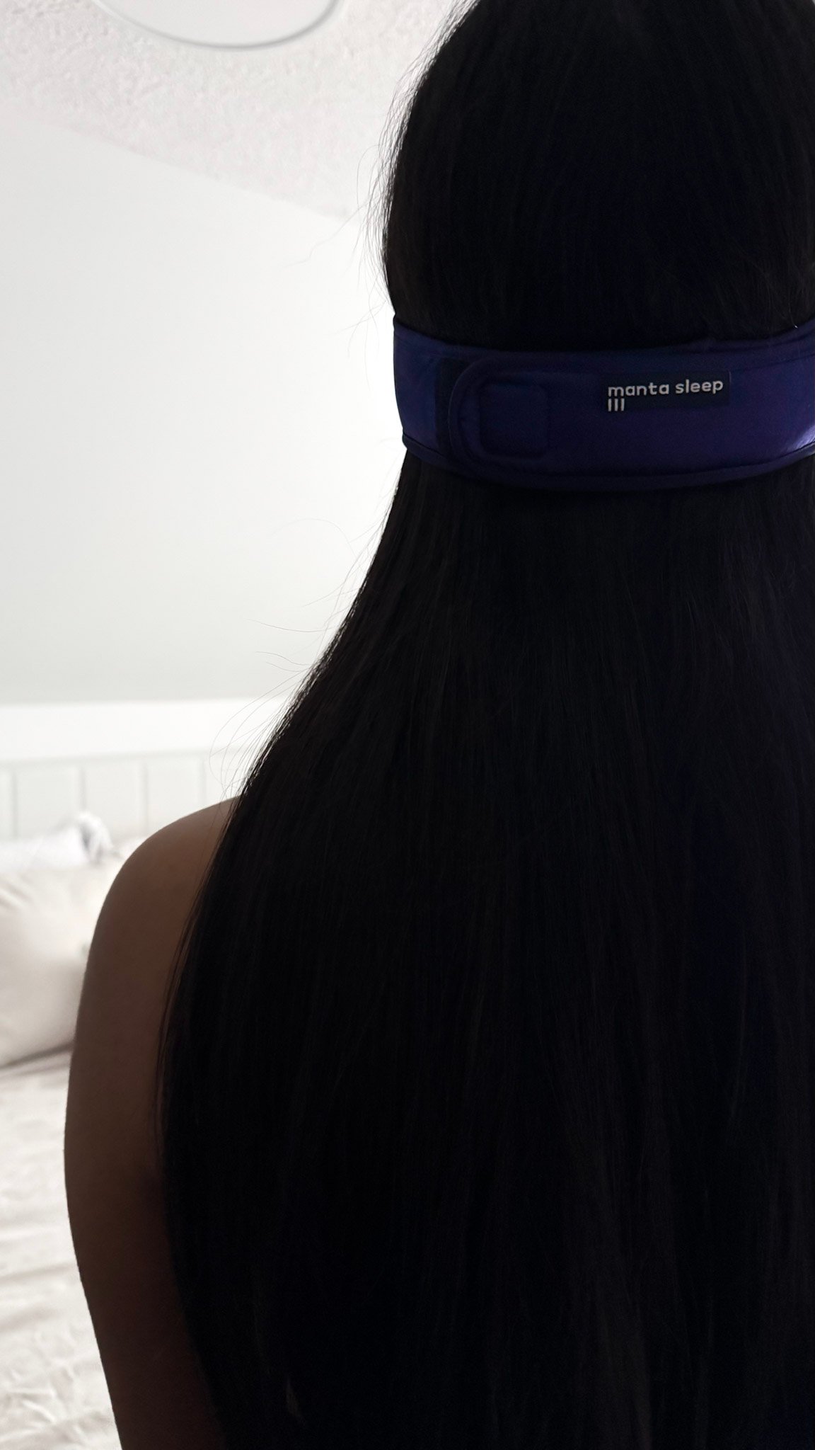 A girl faces away and toward a bed, she is wearing a sleep mask that says Manta Sleep on the band.