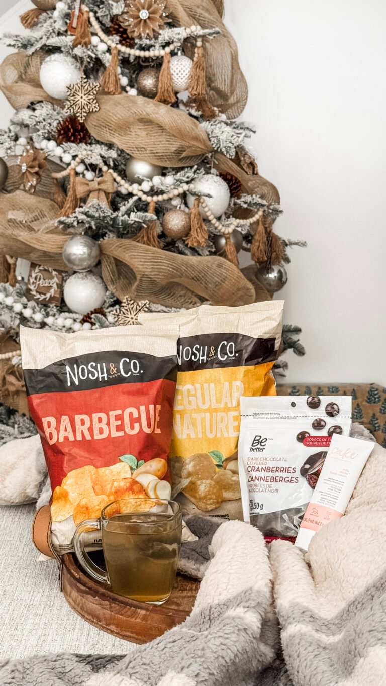 A tray is filed with Nosh & Co Chips in Barbecue and Regular, Chocolate Covered Cranberries, and herbal tea. There's a cozy blanket and Cake hand lotion. A Christmas tree is behind it.