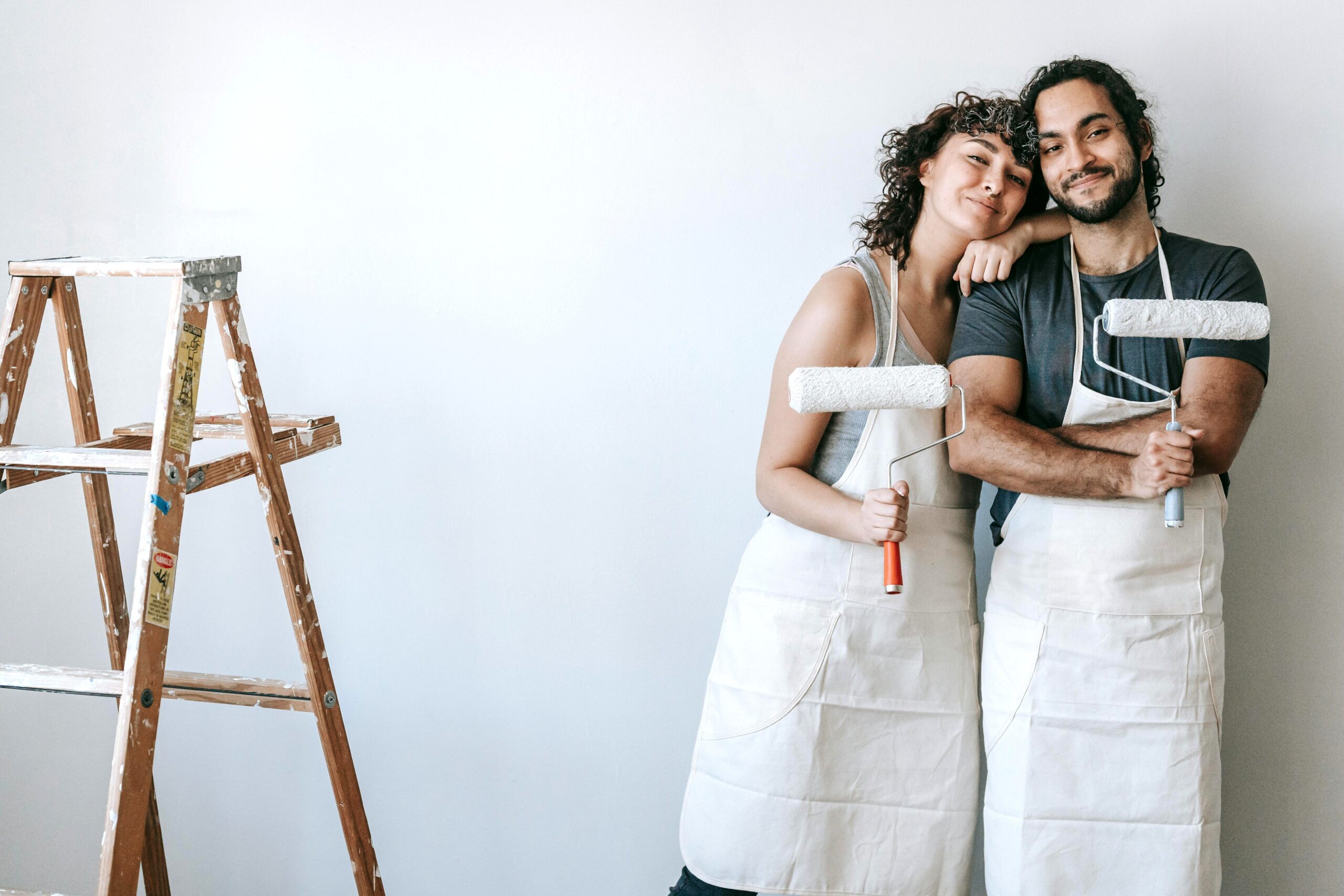 A couple are holding paint rollers are next to a ladder. They are posing together. This article covers home improvement tips for long-term use.