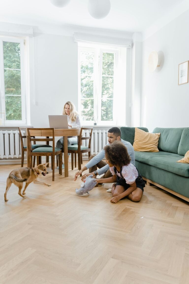 A family relaxes in a living room on the floor with a puppy. The mom works in the back at a dining table. This article covers the advantages of choosing sustainable hardwood floor.