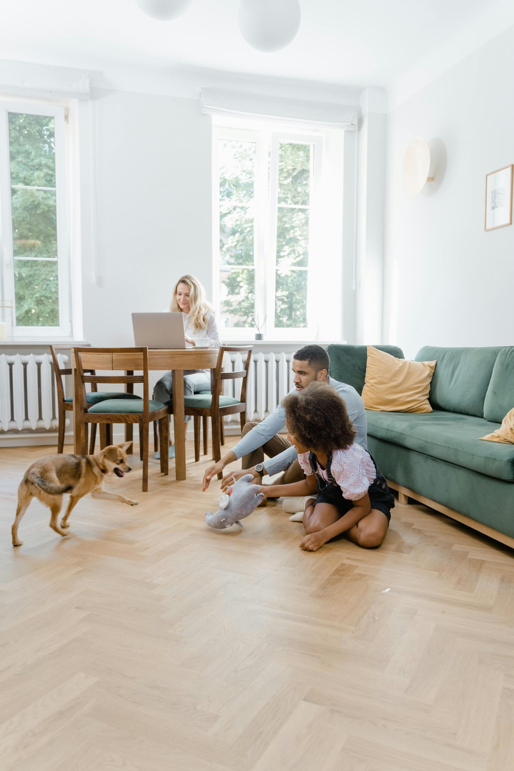 A family relaxes in a living room on the floor with a puppy. The mom works in the back at a dining table. This article covers the advantages of choosing sustainable hardwood floor.