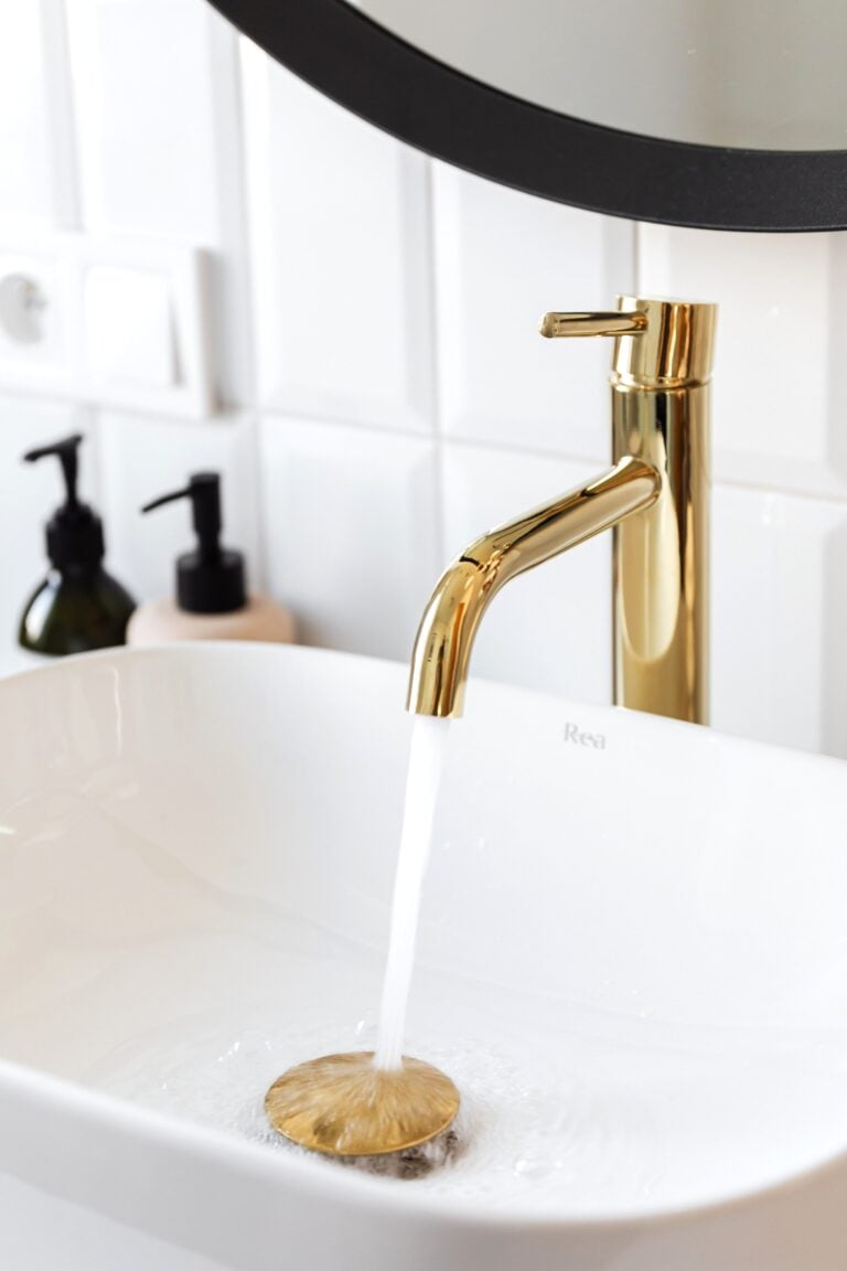 Gold faucet pouring water into a sleek modern sink with tile background. This article shares easy ways to prevent costly plumbing surprises.