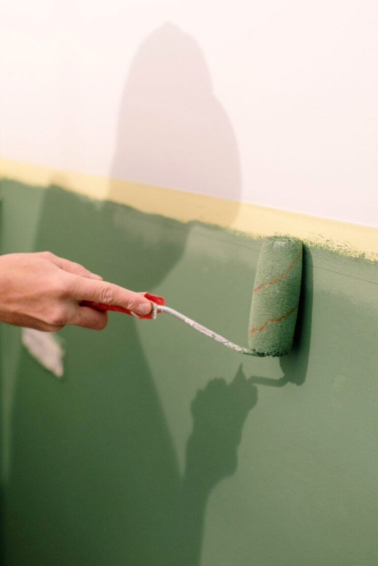 A person using a green paint roller to paint a wall indoors. This article shares how weather affects painting projects.