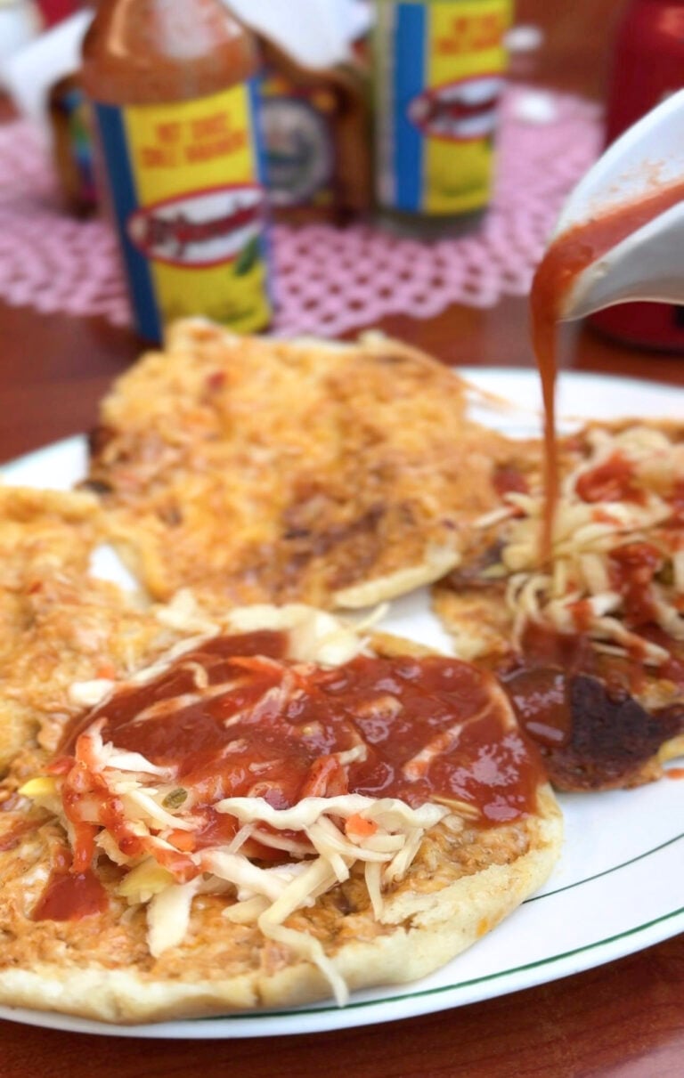 A plate of opened pupusas mixtas sit on a plate, topped with curtido, and being poured over with salsa. This article covers the best pupusas in Windsor.