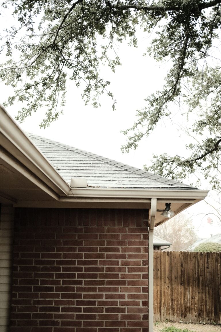 A serene view of a red brick house corner with overhanging tree branches. This article shares the ultimate season-by-season roof care guide.