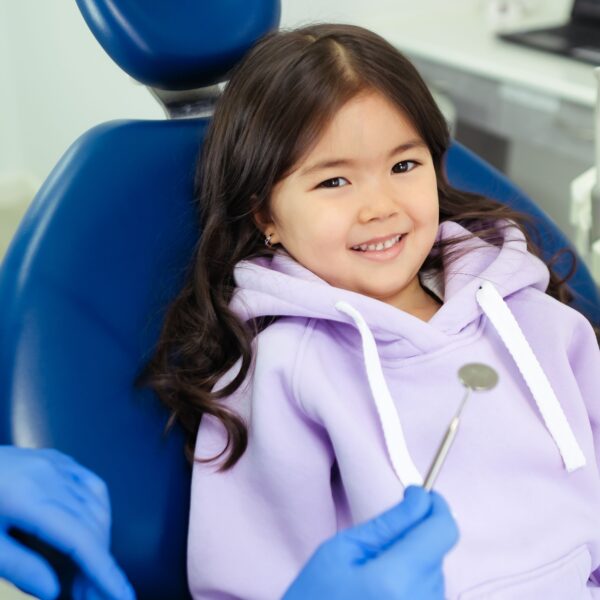 A young girl smiling while sitting in a dental chair, ready for her dental check-up. This article shares reasons families are choosing dental plans for everyday oral care.