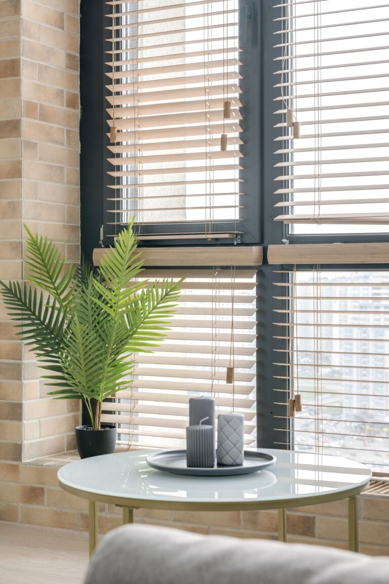 Stylish interior featuring a circular table with candles and a potted plant by large windows with blinds. This article shares what every homeowner should know before their next blinds purchase.