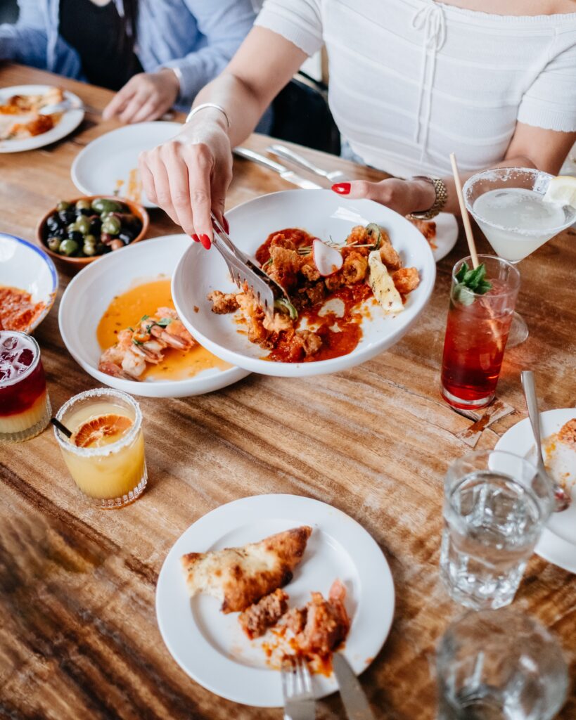 Top view of a dining table with gourmet dishes, drinks, and a woman serving food. This articles helps you plan a great Los Angeles food day.