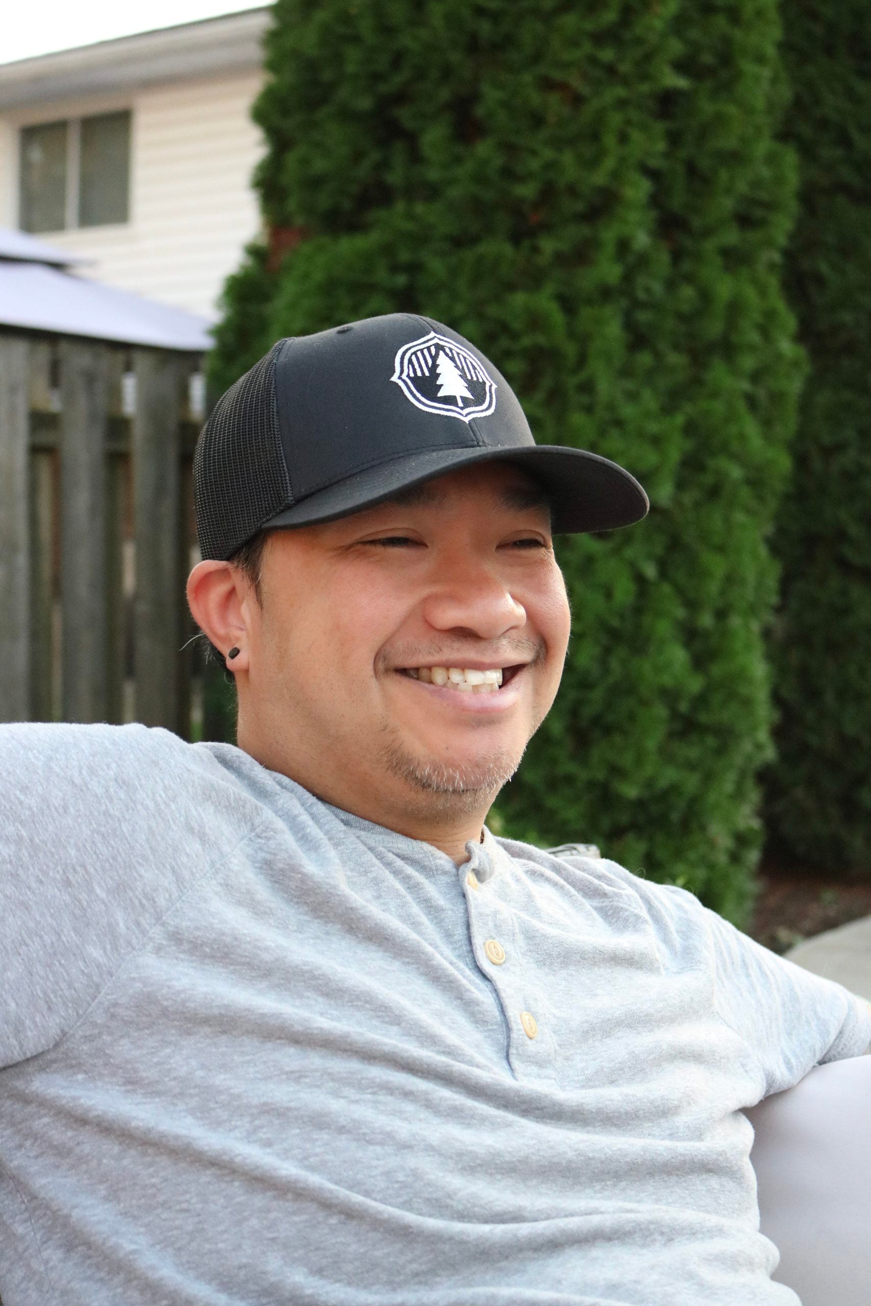 Darasak wears a Timber Stoves hat and smiles while sitting in the backyard patio. The hat is part of the November promo for Timber Stoves.