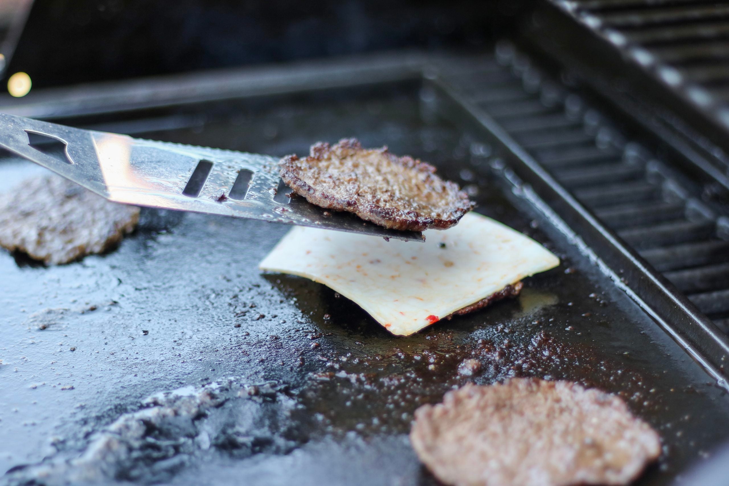 A spatula loads a smash burger on top of another patty with Monterrey jack cheese on a black top on a BBQ. 