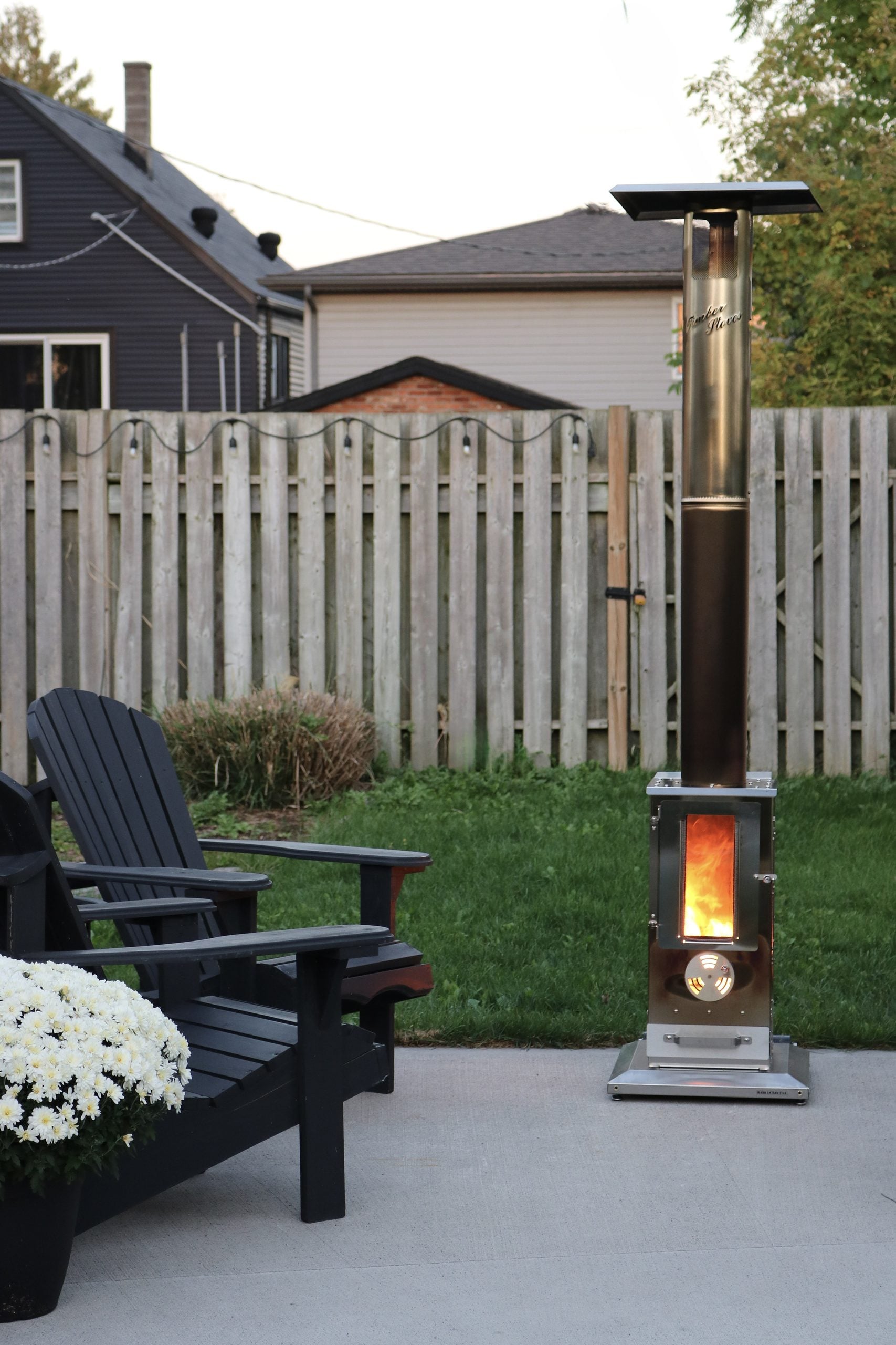 A Timber Stove 'Big Timber' sits on a patio with Adirondack chairs and white mums. This article shares a Big Timber Review and why adding a Big Timber Patio Heater enhanced our backyard all season.