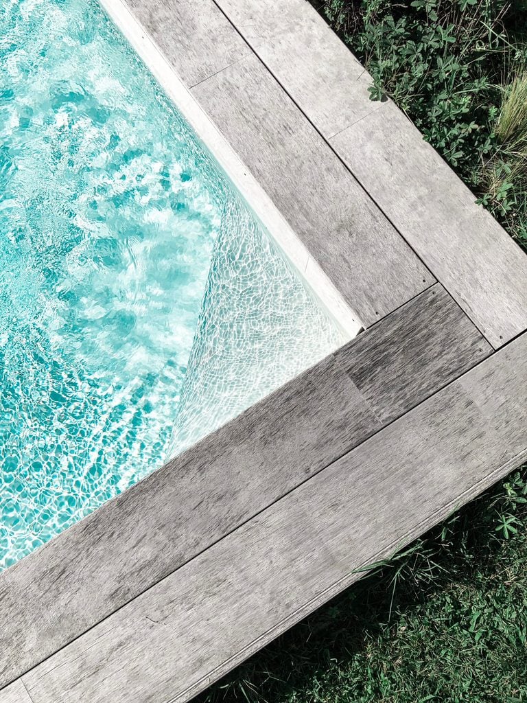 Close-up view of a serene swimming pool with wooden decking surrounded by greenery. This article shares how to create your dream backyard with custom pool construction and design.