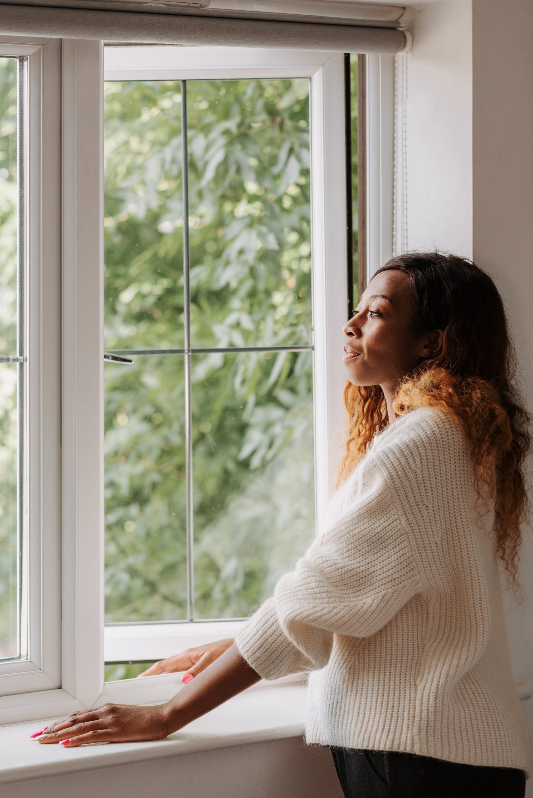 A woman in a white sweater gazes out of a window, enjoying a peaceful moment indoors. This article shares 5 companies that complete a smarter window replacement plan.